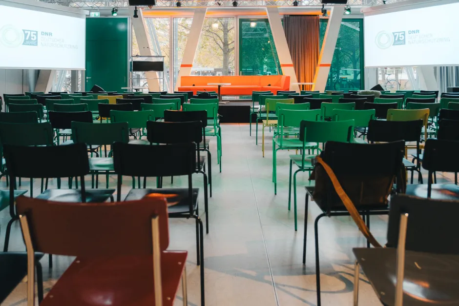 noch leerer Saal in der taz-Kantine mit Blick auf Bestuhlung und Leinwände mit dem 75-Jahre-DNR-Logo